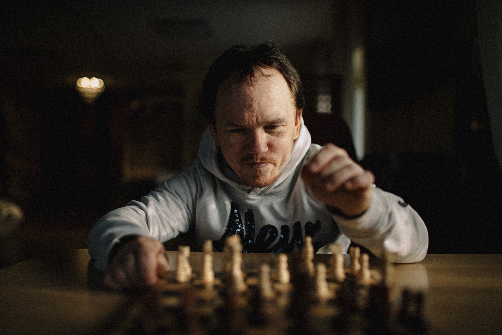 Fotol: Jüri Alandi. Foto autor: Ken Mürk. PVC plaadile prinditud värvifoto. Formaat: A2 rõhtasendis (42x59,40cm). Foto kirjeldus: Mõtliku näoga keskealine, lühikest kasvu mees istub hämaras toas laua taga. Tal on taganeva juuksepiiriga, lühikesed, tumepruunid juuksed. Paaripäevase habeme ja vuntside vahelt paistavad pisut pruntis huuled. Mehe pilk on suunatud tema ees laua peal olevale malelauale. Jüril on seljas helehall kapuutsiga pusa. Suurelt üle rinnaku tumesinistest, helkelvatest litritest inglisekeelne sõna „New“. Jüri parem käsi on toetatud malelaua äärele. Vasaku käe küünarnukk toetub lauale ja töntsakad sõrmed on valmis haarama valgeid malendeid, mis asetsevad temapoolsel mänguväljal. Toa laes kollaka valgusega kuppellamp. Pildil paremalt poolt aknast kumab pisut päevavalgust. Hägustatud ja udune taust.